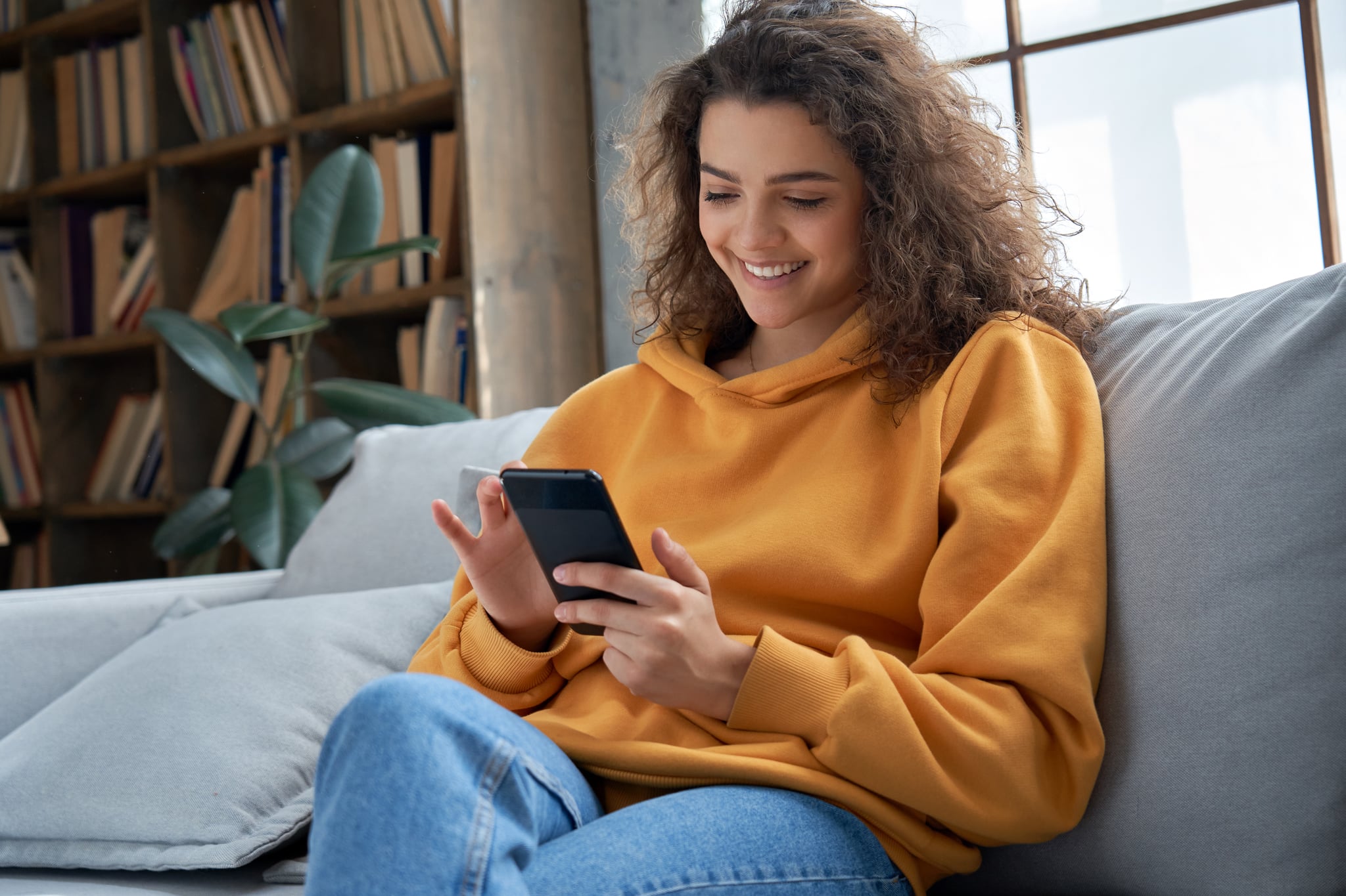 快乐的千禧西班牙少女拿着智能手机在家检查社交媒体。微笑的年轻拉丁女子使用手机应用玩游戏，网上购物，订购快递放松在沙发上。