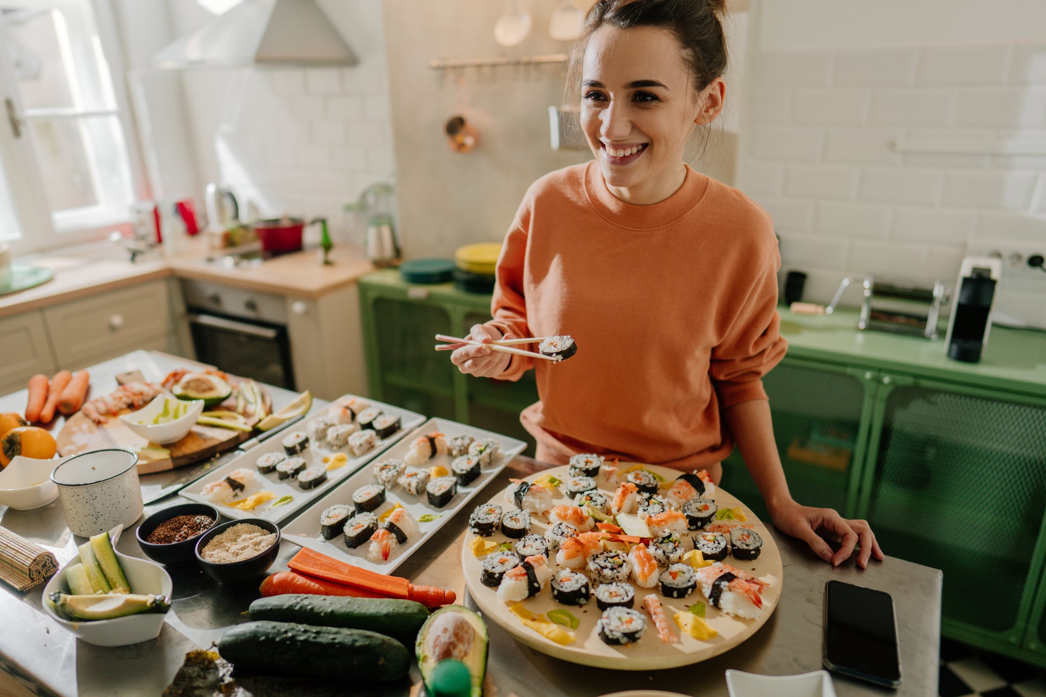 Photo of a smiling young woman holding a sushi roll with a pair of chopsticks by the kitchen counter.