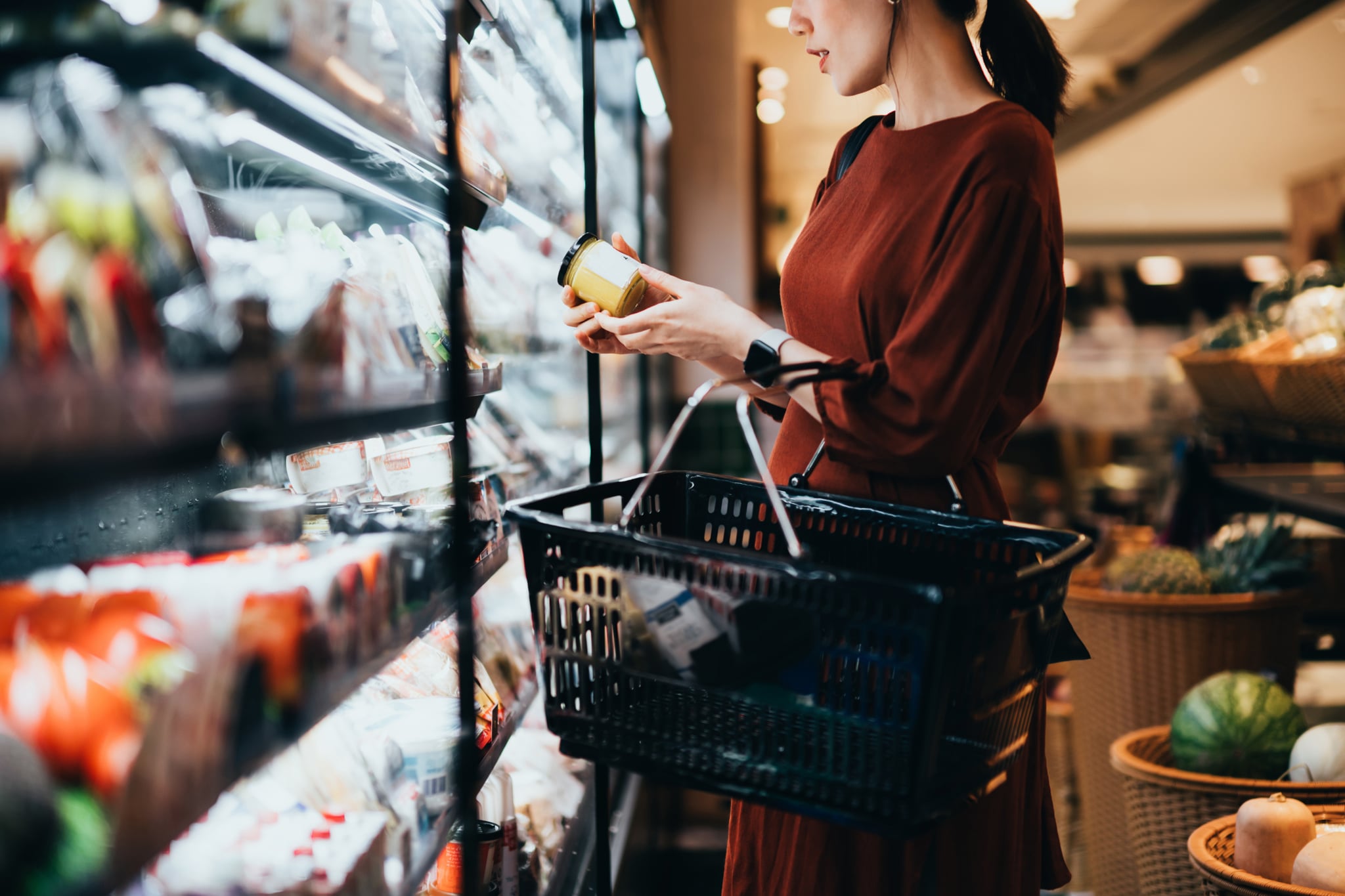 年轻的亚洲妇女拿着一个购物篮，站在乳制品通道，阅读营养标签上的新鲜有机健康酸奶。选择更健康的食物