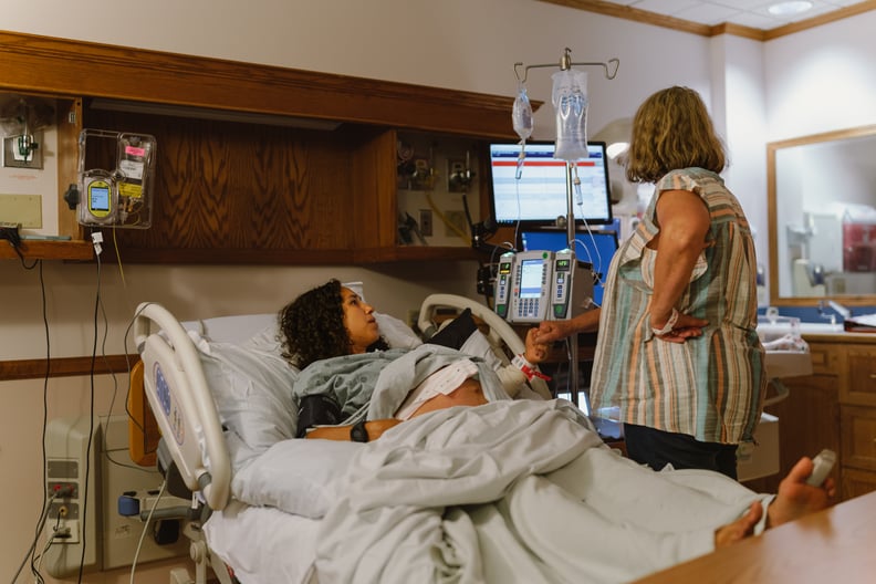 A pregnant Latine woman lies in a hospital bed and holds a caregiver's hand while having labor contractions.