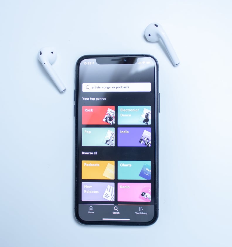 A pair of AirPods is shown next to an iPhone displaying the Spotify search bar, top genres, and different features of the app such as Podcasts and Radio.