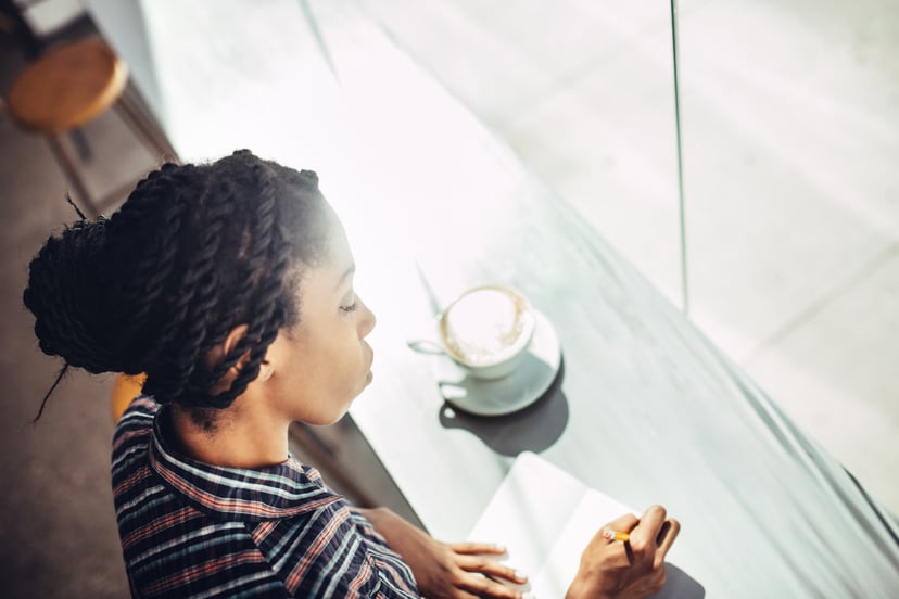 A beautiful young African American woman sits at the window counter of a coffee shop, enjoying a latte while writing ideas down in a small notepad journal or diary.  She wears a casual button up shirt, with her hair up, and a content relaxed look on her f