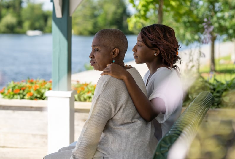 A young woman sits outside beside her Mother, with her arm around her and one hand on her shoulder. They are looking out over the water as she tries to comfort and support her Mother through her cancer diagnosis. They are both dressed casually as they spe