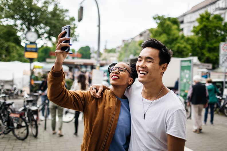 A young tourist couple standing in a new city take a photo of themselves using a smartphone.