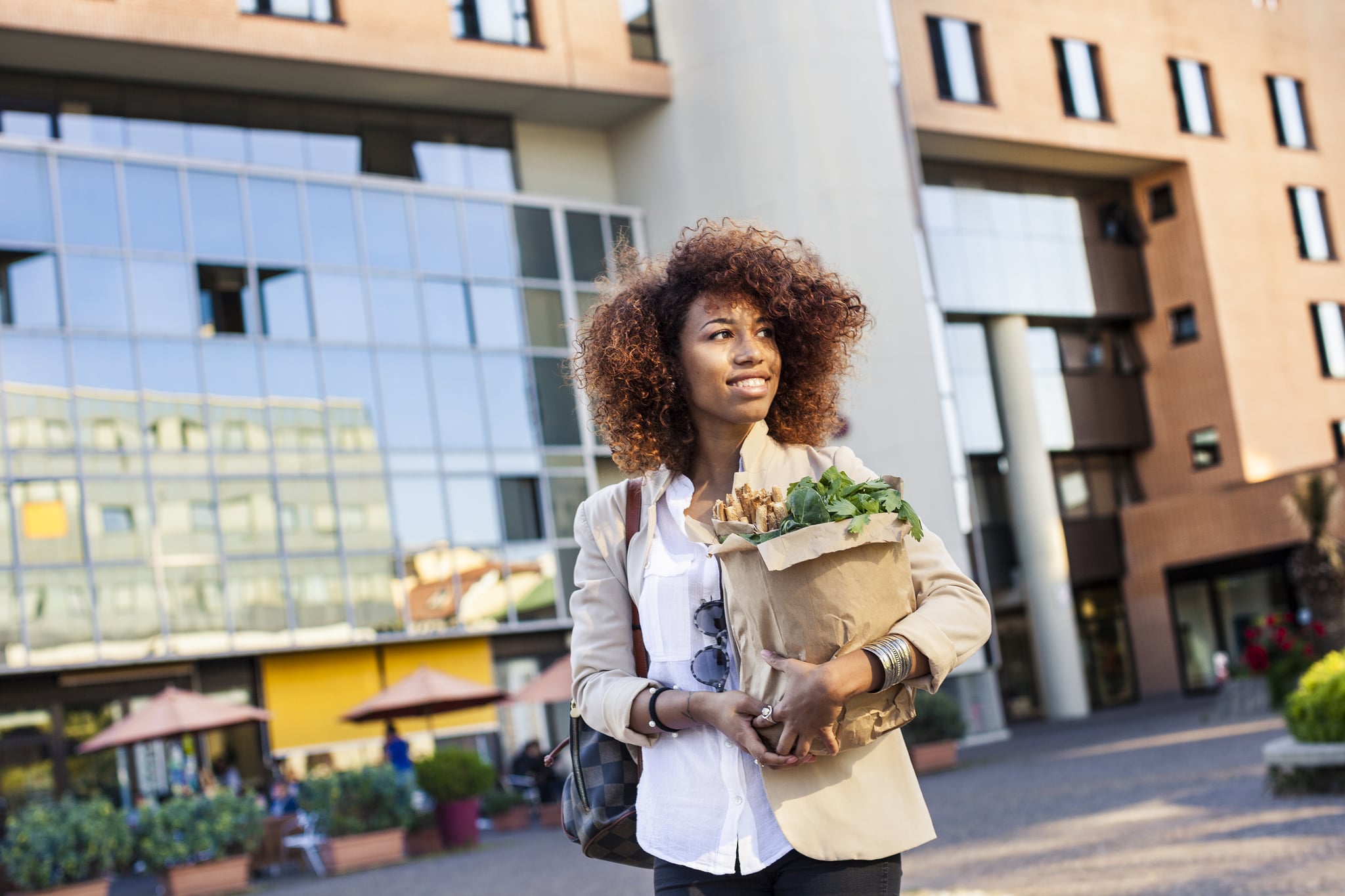 Young afro woman holding a shopping bag full of green vegetables and sesame breadsticks walking in the morning before going at work.