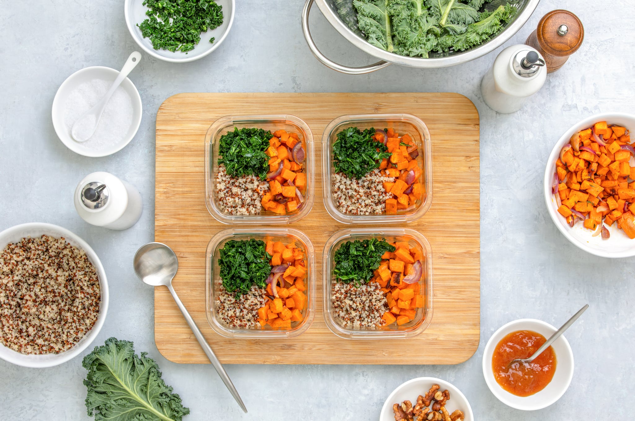 Quinoa, kale and roasted sweet potato lunch boxes are standing on a kitchen table with ingredients for cooking, overhead view, fall healthy take-away lunch recipe concept