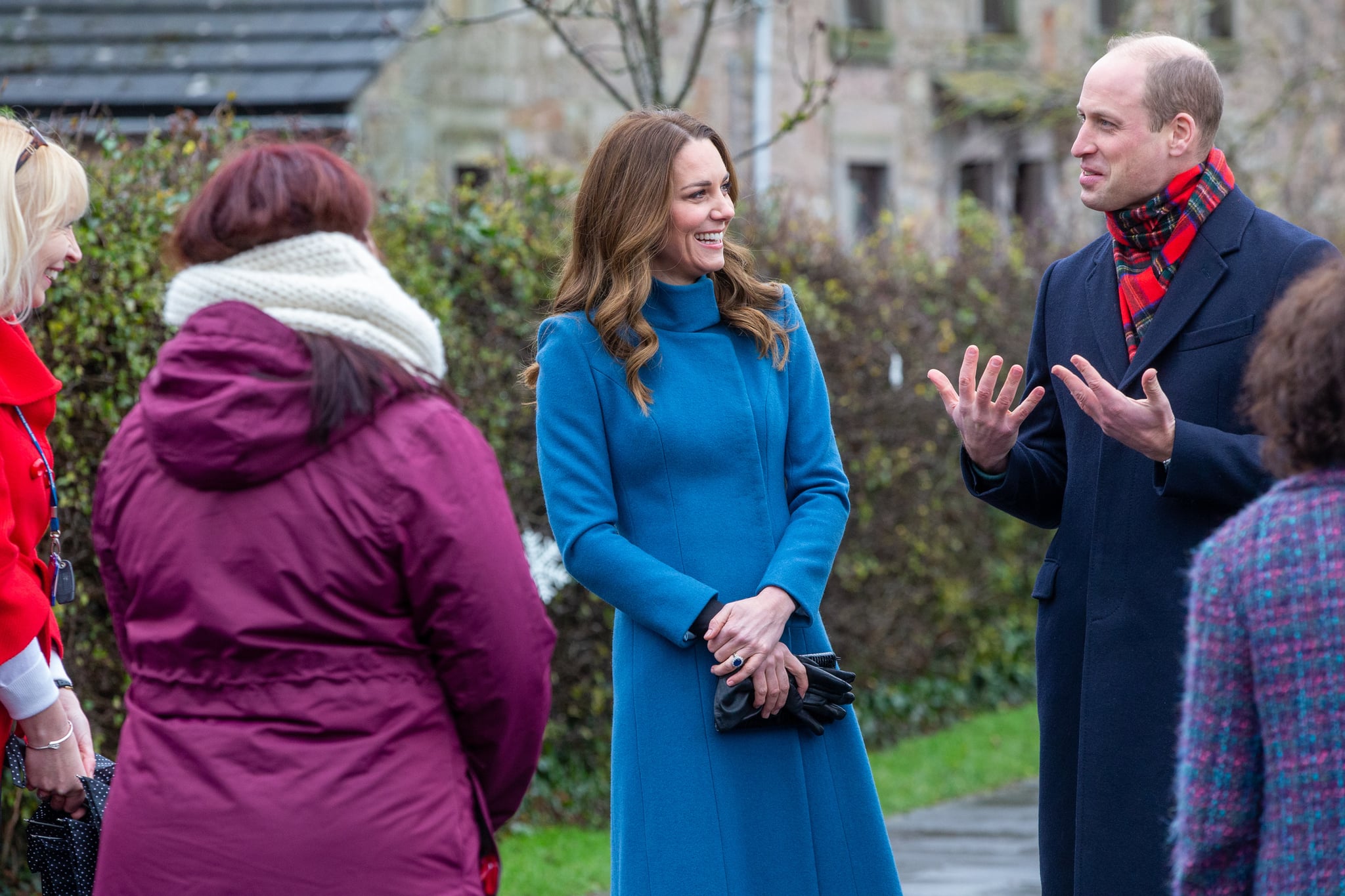 Kate sorri enquanto olha para William que gesticula ao ar livre. Eles ainda estão com as roupas dos compromissos na Escócia. Há três professoras de costas.