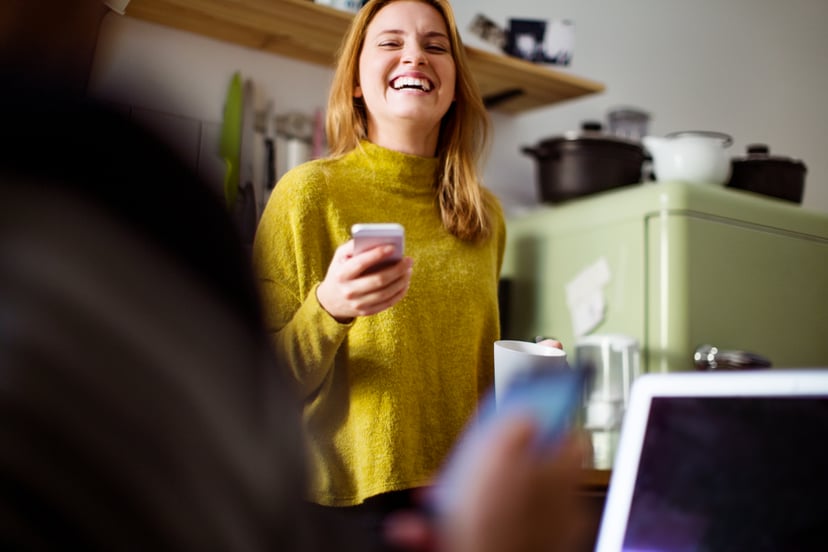 Young woman talking to man sitting in front and smiling. Cheerful female with mobile phone in kitchen.
