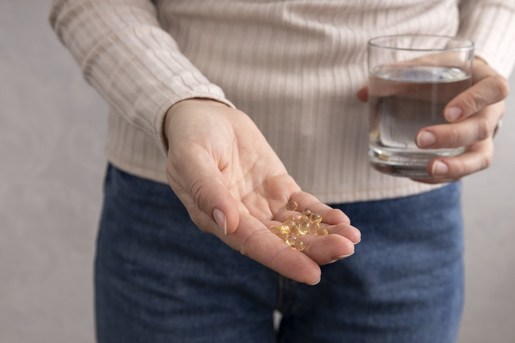 Closeup woman holding vitamin D capsules in one hand and a glass of water in the other while standing against bright wall.