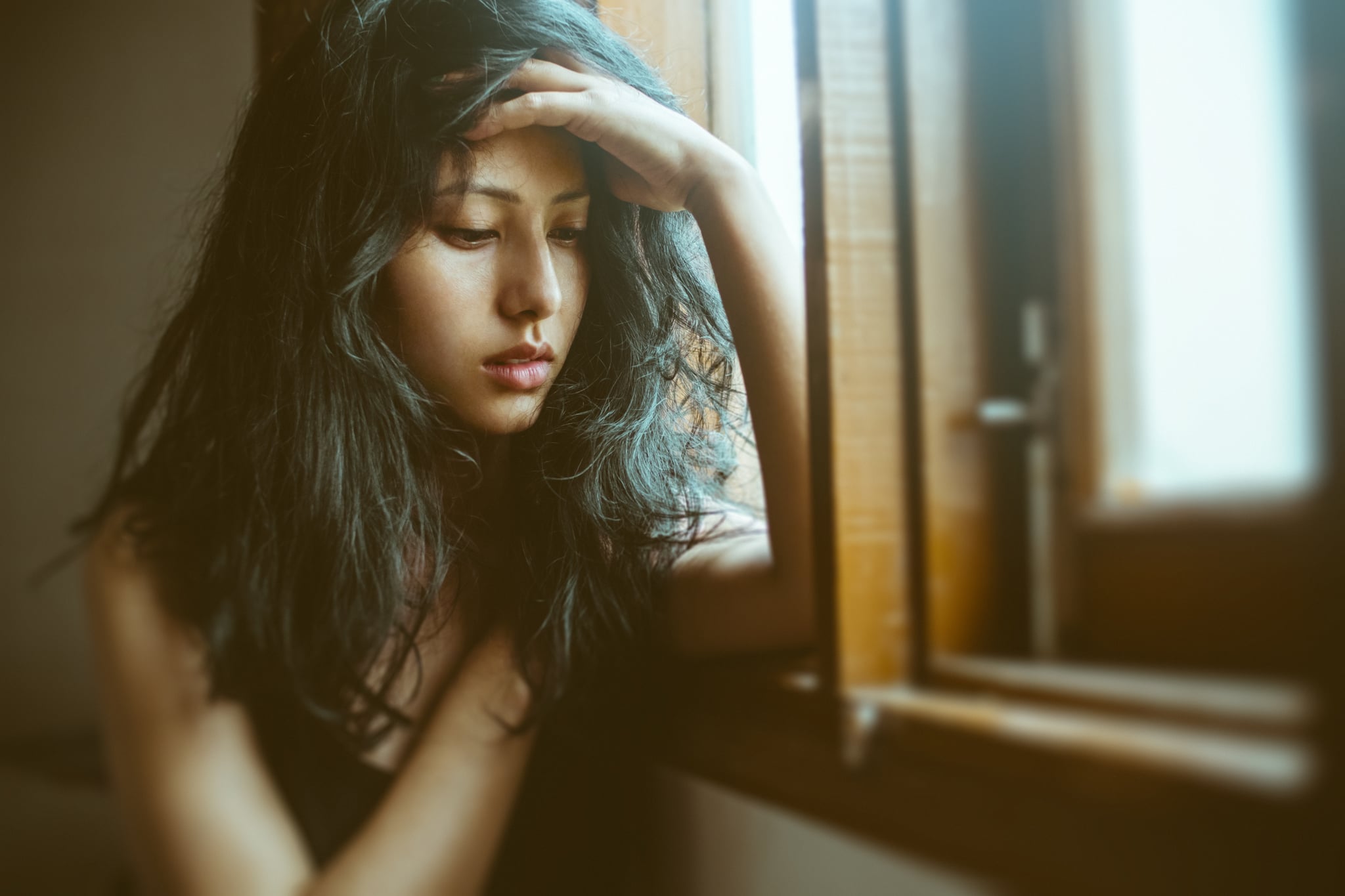 Portrait of Asian, Indian sad, serene young woman sits near window at home. She looks down, holds her head and thinks deeply with blank expression.