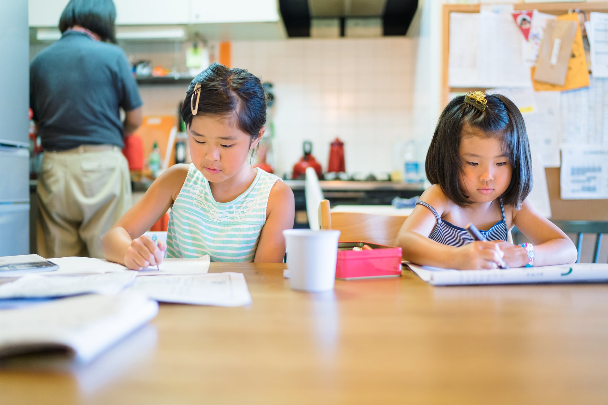 Two siblings are doing their homework while their father is cleaning the kitchen in the background.