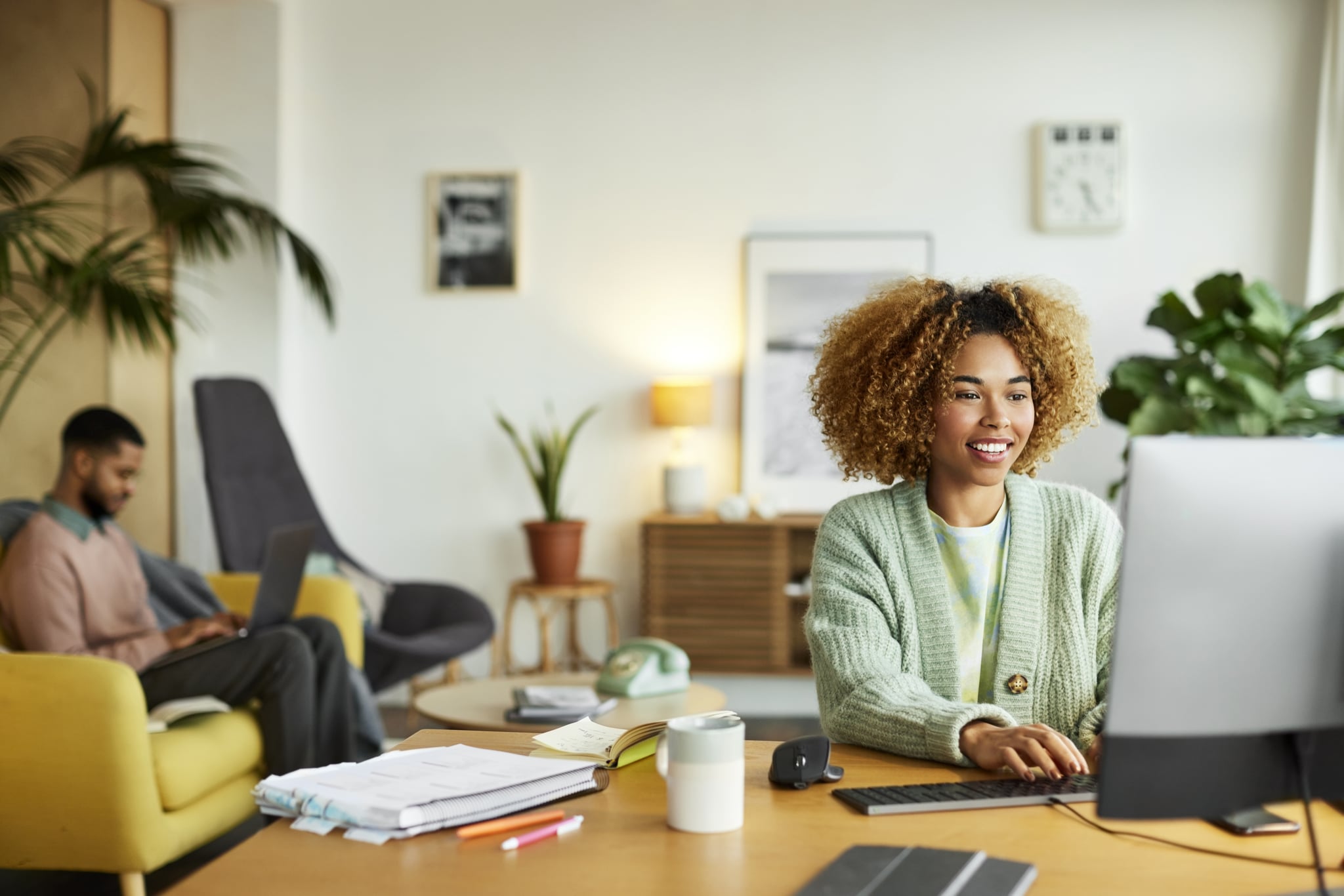 Smiling female freelancer using computer while colleague in background. Businesswoman is working at desk in home office. They are in apartment.