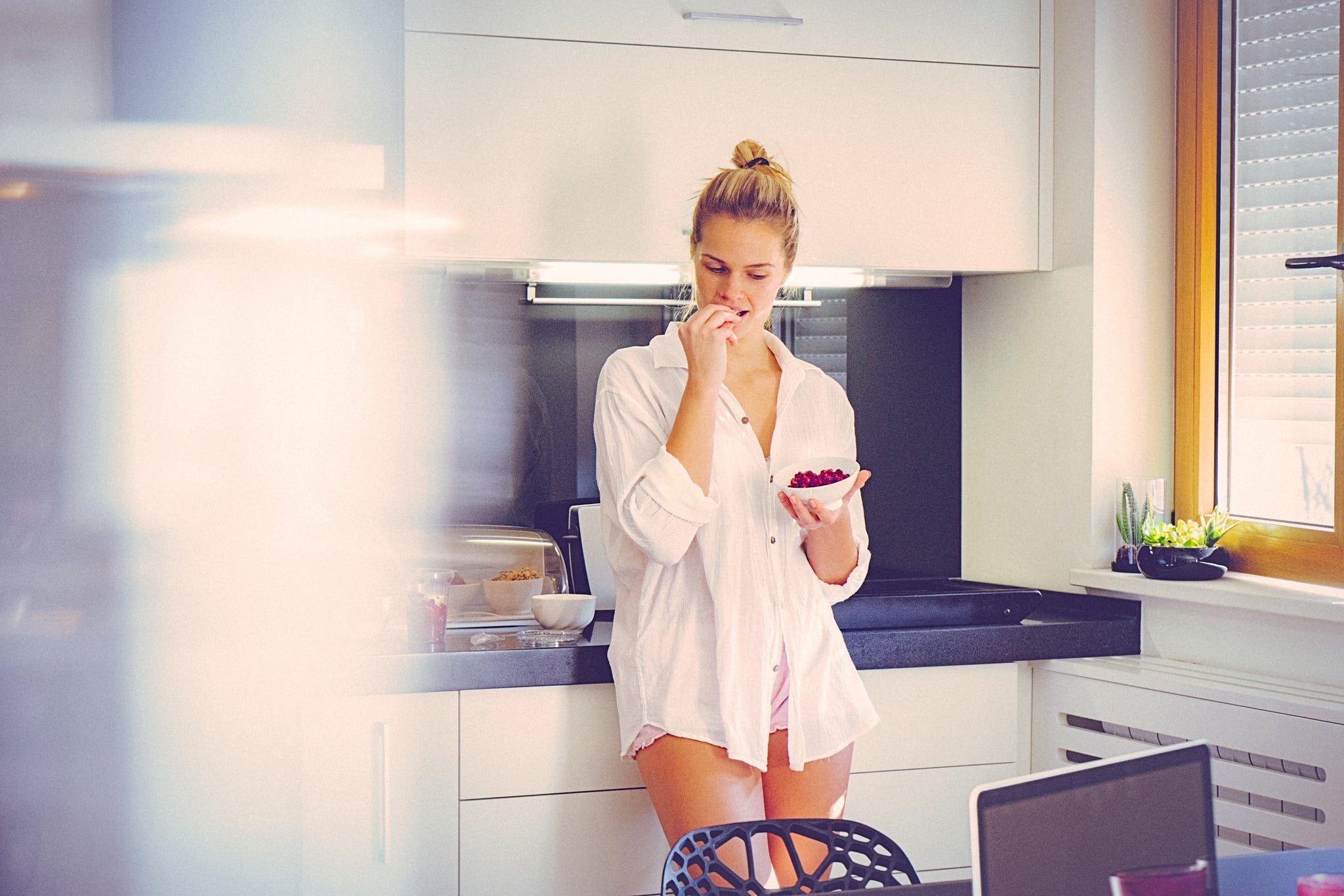 Attractive blonde woman in the morning, at home in the kitchen, eating pomegranate. The woman is dressed in pajamas and it seems that she recently got out of the bed and started to prepare breakfast. Her long blonde hair is swept back from her face. She seems sleepy. The shot is executed with available natural light, and the copy space has been left. Shallow DOF. Soft focussed.