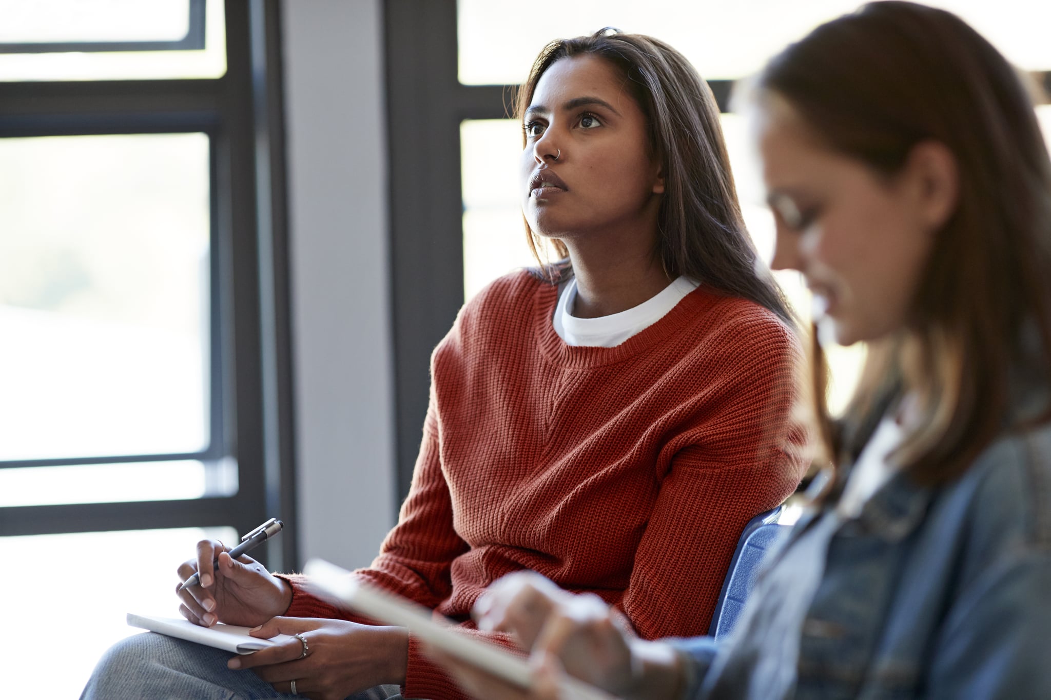 Thoughtful young woman looking away while sitting by female friend using digital tablet in classroom at community college