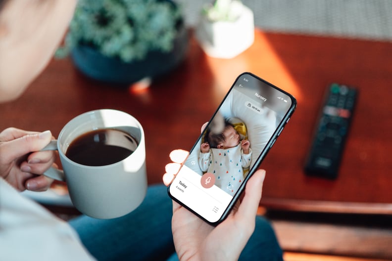 Over the shoulder view of young mother monitoring her baby girl with smart home app. Smart baby CCTV. Using baby smart monitor app on mobile phone. Smart home camera.