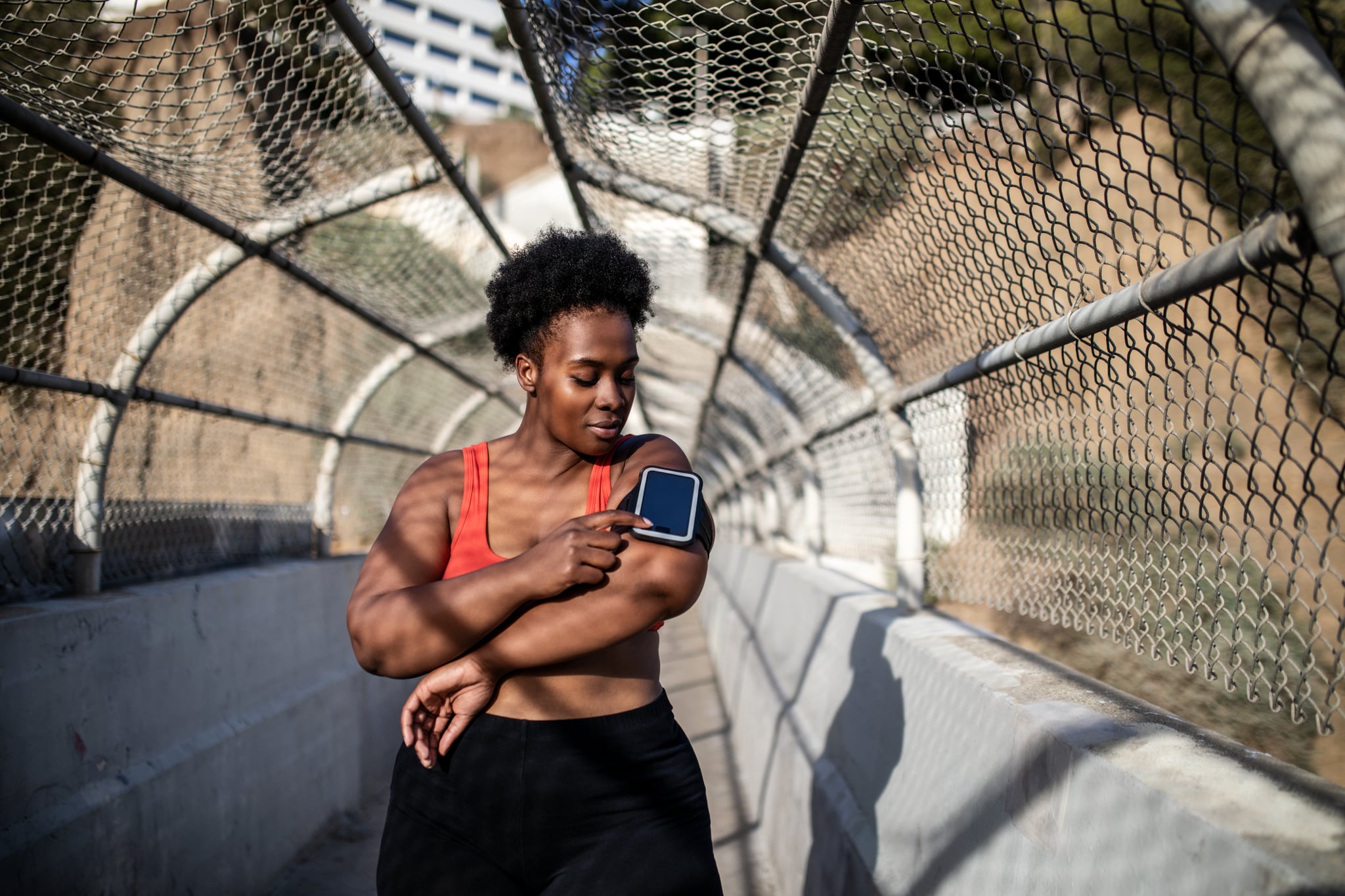 Healthy woman touching phone screen on armband before exercising outdoors. African american woman in sports clothing using phone while exercising outdoors.