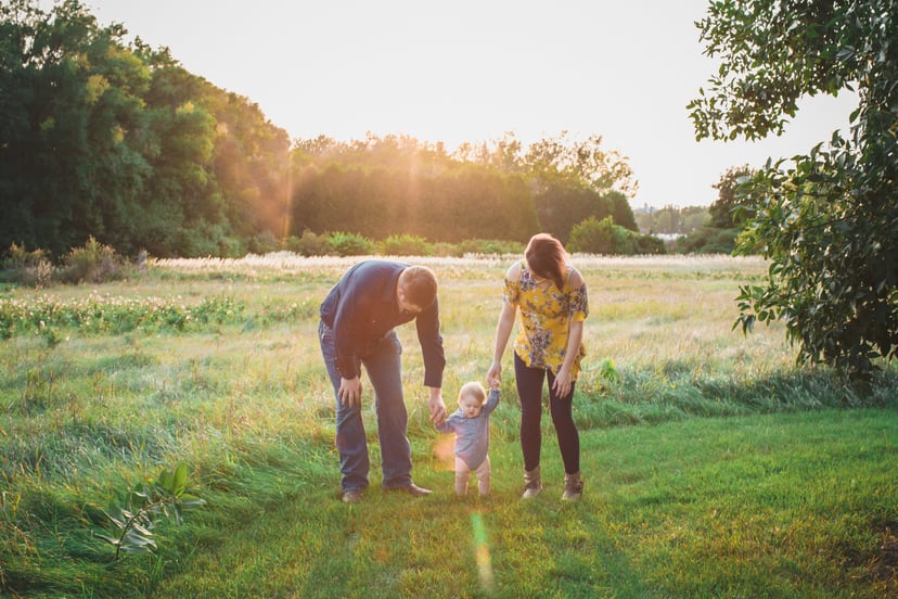 Mother and father stand on either side of their baby boy and hold his hands and try to help him walk across a grassy meadow.