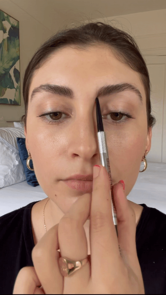 First, you brush the hair in your brows up. Then, holding the brow pencil vertically, you use the outer corner of your nose to mark the inner bottom corner of your eyebrow on each side — this marks the start of your brows. Next, you line the pencil up at an angle extending from the corner of your nose to the end of your brow and mark the placement.
