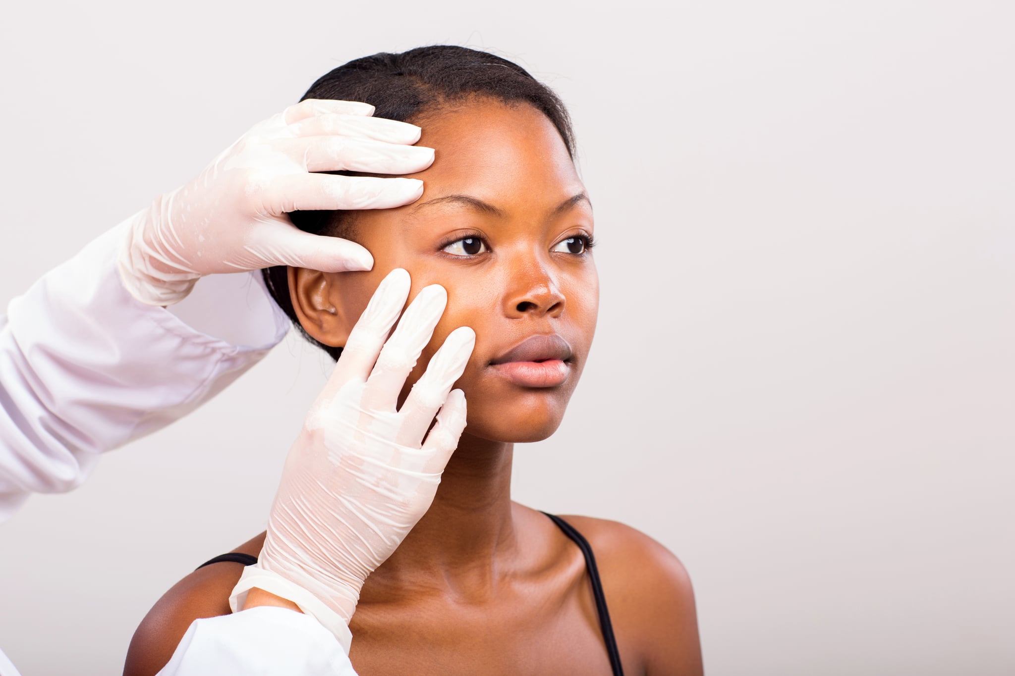 dermatologist checking young african american woman face skin on plain background