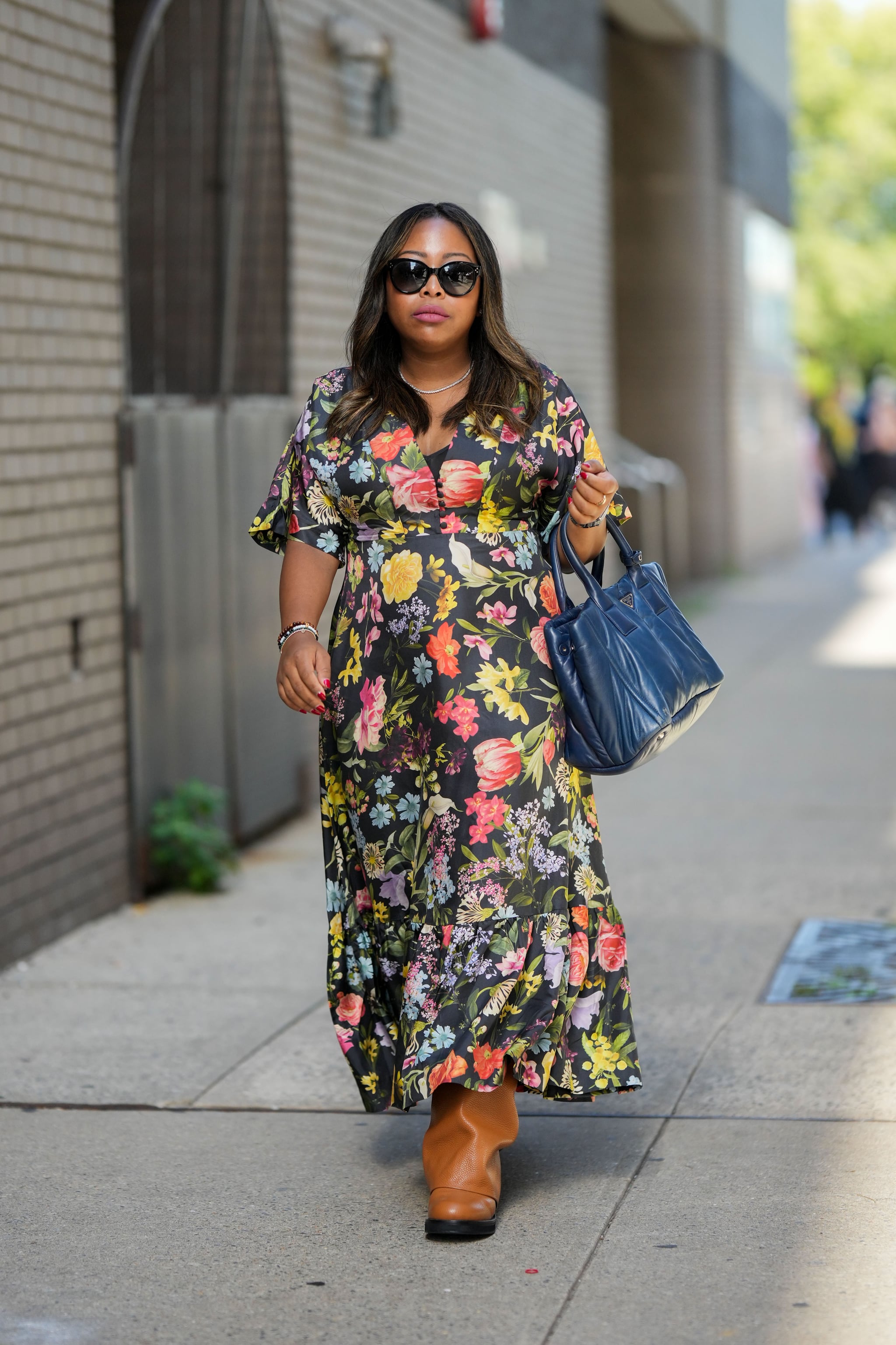 NUEVA YORK, NUEVA YORK - 10 DE SEPTIEMBRE: Un invitado usa gafas de sol negras, un collar de diamantes, un bolso grande de cuero azul marino brillante de Prada, anillos de oro, un reloj plateado, pulseras, un vestido negro con estampado de flores multicolores mangas cortas / vestido largo , botas hasta la rodilla de cuero marrón brillante, en las afueras de Altuzarra, durante la Semana de la Moda de Nueva York, el 10 de septiembre de 2022 en la ciudad de Nueva York.  (Foto de Edward Berthelot/)