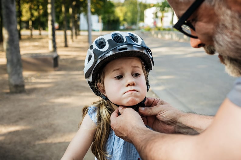 A grandpa carefully doing up his granddaughters crash helmet while on a day out at the park together.