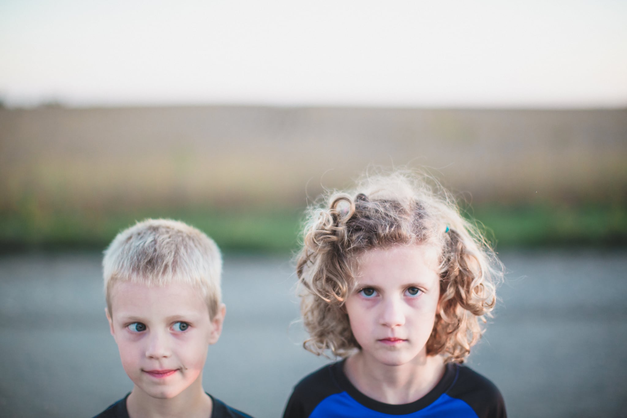 Close up portrait of a boy and girl standing side by side. Girl with curly hair looks sternly into the camera while blond boy looks at her with side eyes and a mischievous expression.