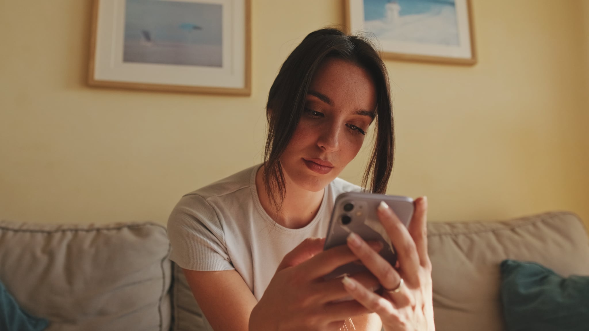 Young woman using mobile phone for texting and scrolling while sitting on sofa at home