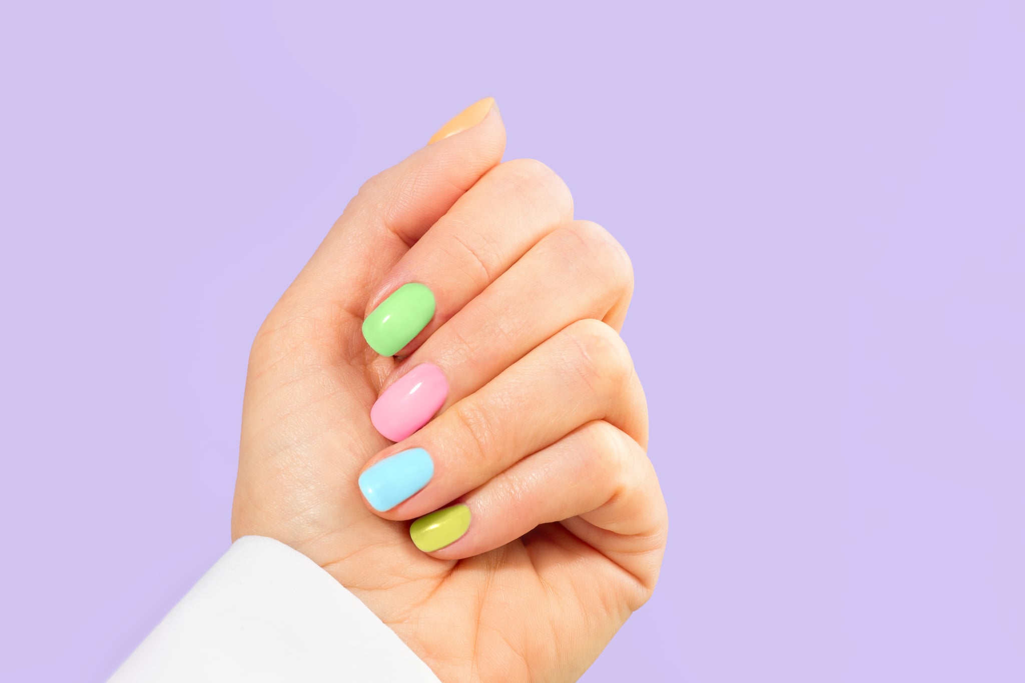 Elegant woman's hand with multi-color pastel manicure on Easter Holiday. Stylish trendy manicure.  Woman dressed in fashionable white shirt. Trendy Color of the Year 2022 Very Peri purple violet lavender color background.  Luxurious background design. Concept of  Easter, Valentine's day. Front view. Copy space. Close-up.