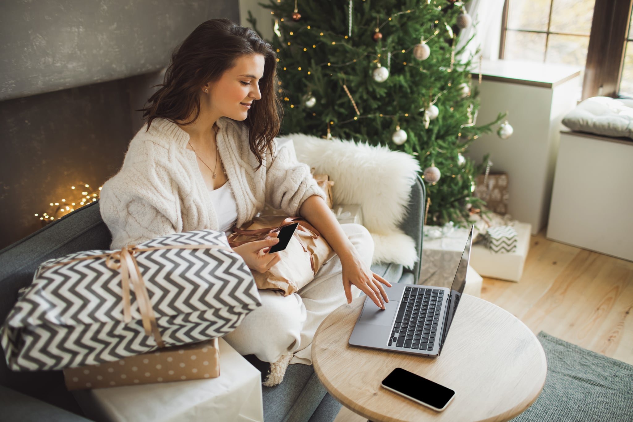 Young happy woman sitting on blue sofa and  holding credit card against the background of Christmas decor and present. Christmas and New Year shopping on Internet, payment by credit card.