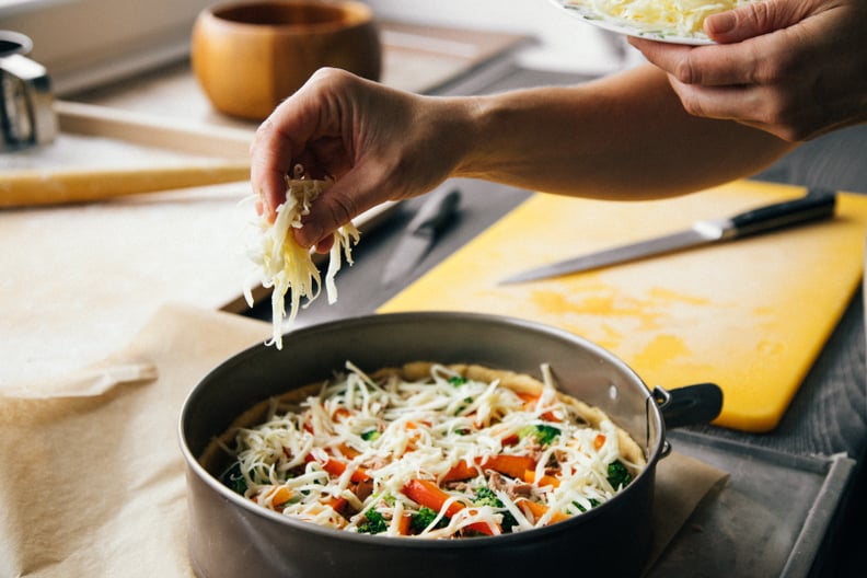Hands of female baker adding grounded cheese on top of uncooked pie stuffed with tuna, broccoli and red bell pepper.