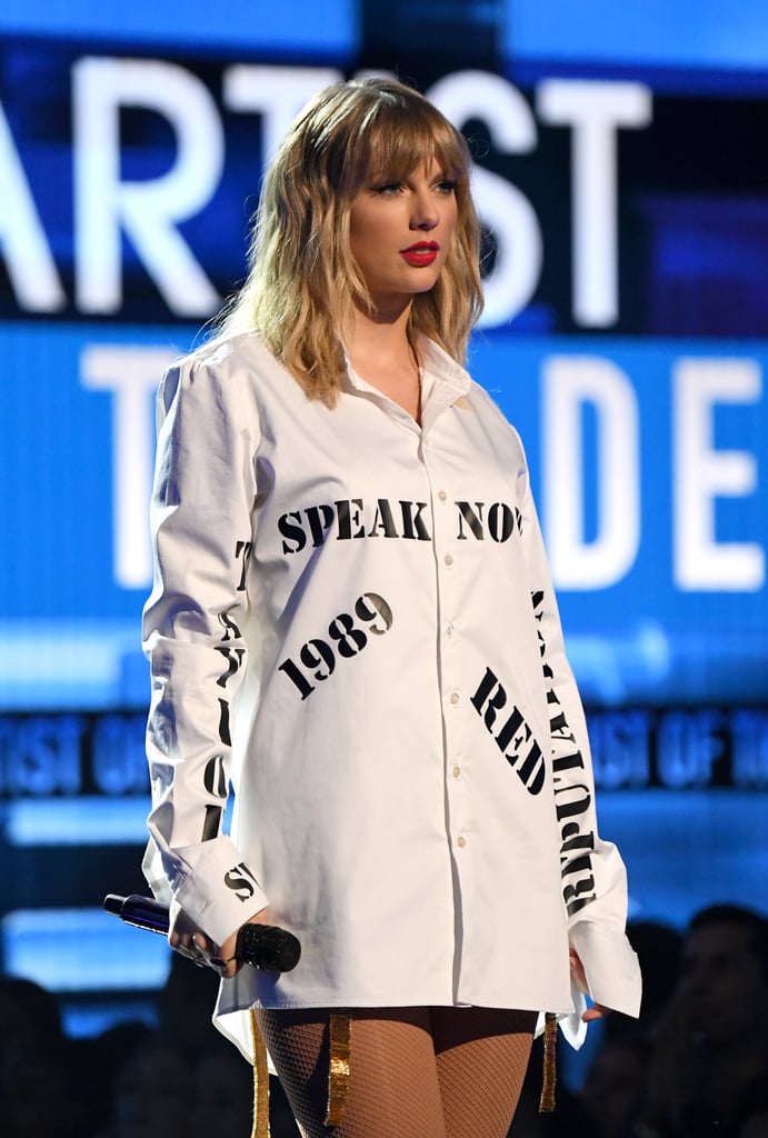 Taylor Swift didn't just rule the red carpet at the American Music Awards, she also used her highly anticipated performance to make a powerful statement about her music. The singer has had an ongoing feud with Scooter Braun and Scott Borchetta regarding the rights to her first six albums, which originally left a question mark hanging over whether Taylor would actually be able to perform her past hits during the AMAs. A compromise was eventually reached, but that didn't stop Taylor from making a pointed message with her wardrobe. 
Taylor stepped out on stage wearing an oversize white shirt bearing the names of her albums Speak Now, 1989, Red, Fearless, and Reputation, printed in a prison-like font, perhaps suggesting that she feels like people have held her prisoner with her own work. The choice of song for this moment, "The Man" from her new album Lover, was also a poignant choice. However, the shirt was eventually removed to reveal a golden beaded bodysuit underneath, as Taylor became a glistening golden goddess as she performed more of her most famous hits, including a memorable version of "Shake It Off" with her friends Camila Cabello and Halsey. Keep reading to see both stage looks ahead.