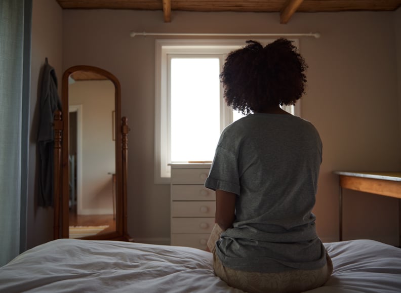 Rear view of a young Black woman sitting on her bed and looking out the window.