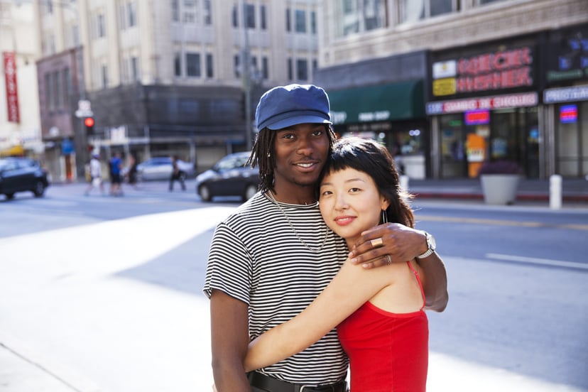 a young couple showing affection while looking at the camera and smiling with a city street scene in the background