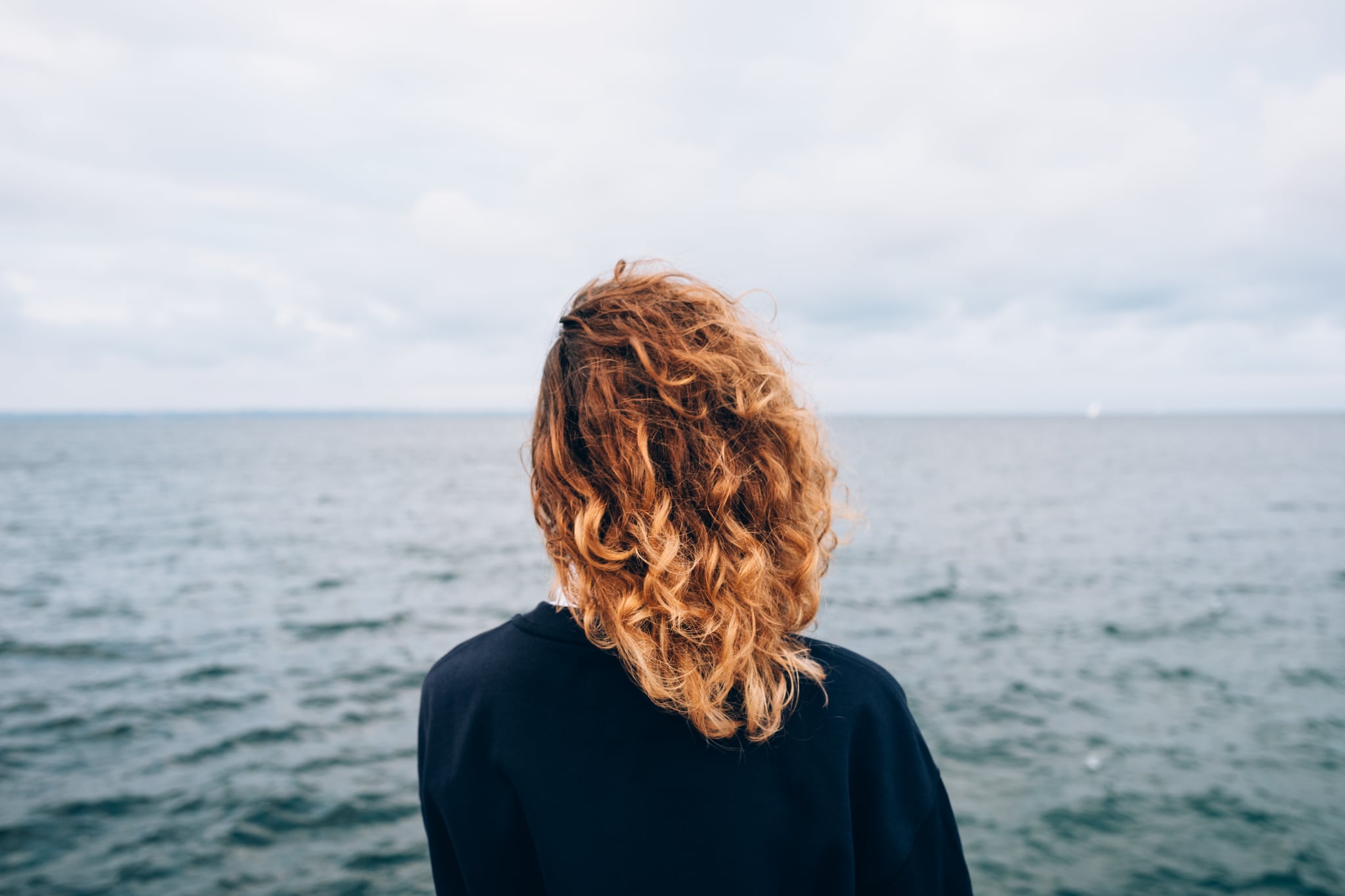Female with red curly hair outdoors standing alone thinking near blue water. Rear view of young woman looking at sea.