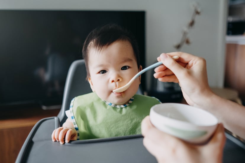 Mother's hand feeding baby girl food on high chair at home