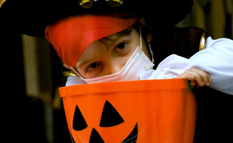 Sad little boy wearing a halloween pirate costume and a protective face mask looking through a window holding a Jack o Lantern bucket