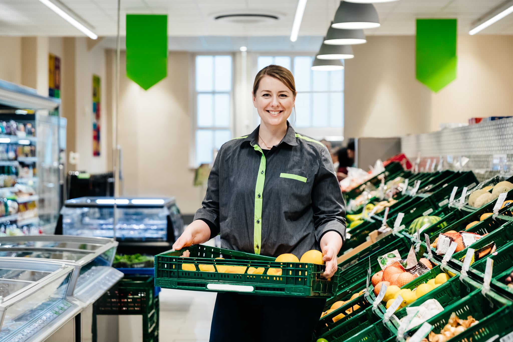 A portrait of a supermarket employee smiling and holding a crate of fruit.