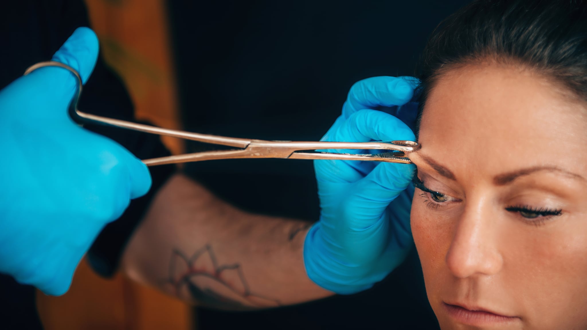 Beautiful young woman getting her eyebrow pierced, body piercer wearing blue gloves