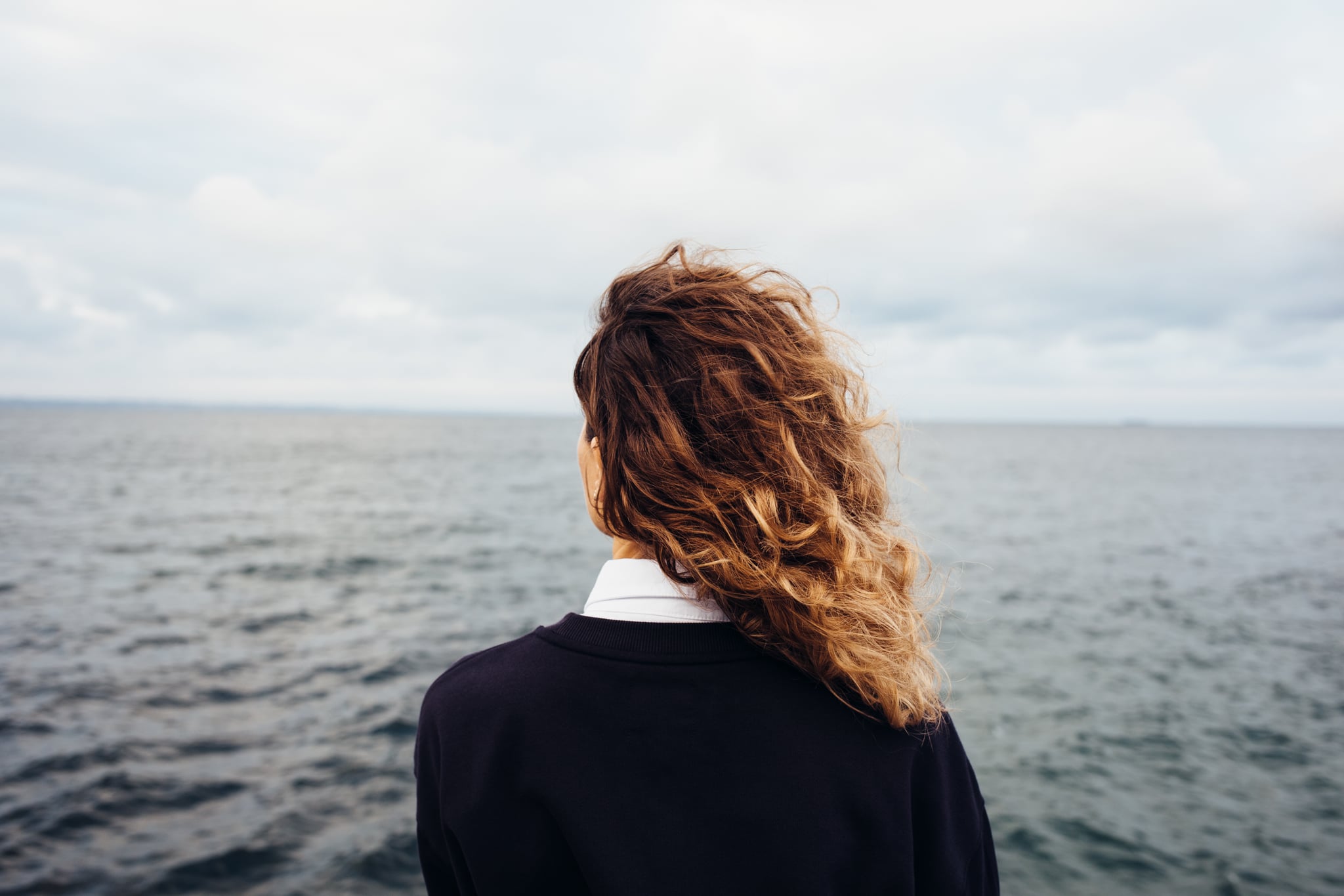 Rear view of young woman looking at overcast sky and grey sea. Female with red curly hair standing alone thinking at the background of seascape.