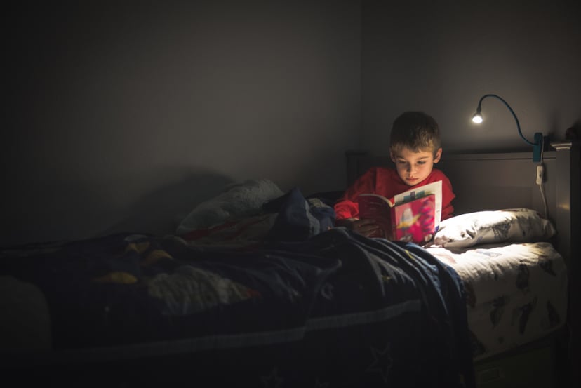 An 8 year old boy is in his bed at night.  His room is dark and lit only by his reading lamp illuminating him and his book.
