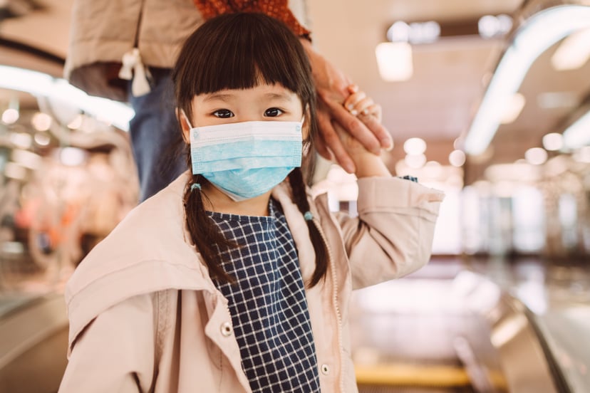 Lovely little girl with medical face mask holding her mom's hand, looking at the camera while standing on the ascending escalator in the shopping mall