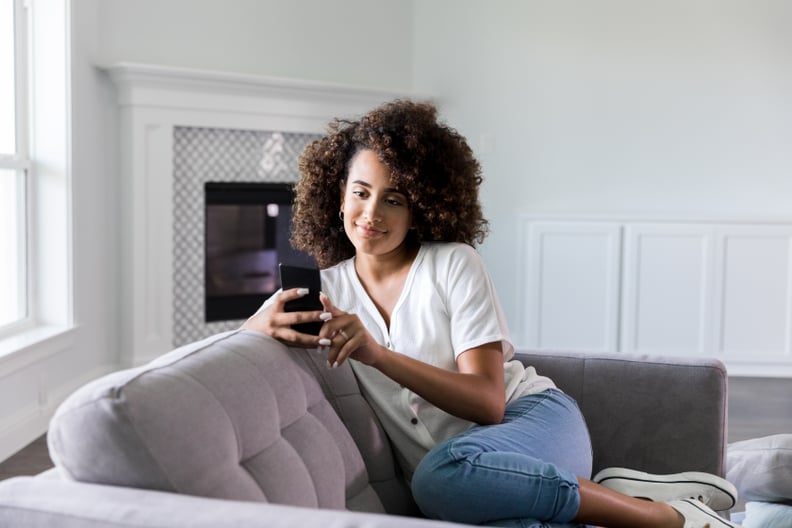 Relaxed young woman smiles while video chatting with friend on smartphone. She is relaxing on the sofa in her living room.