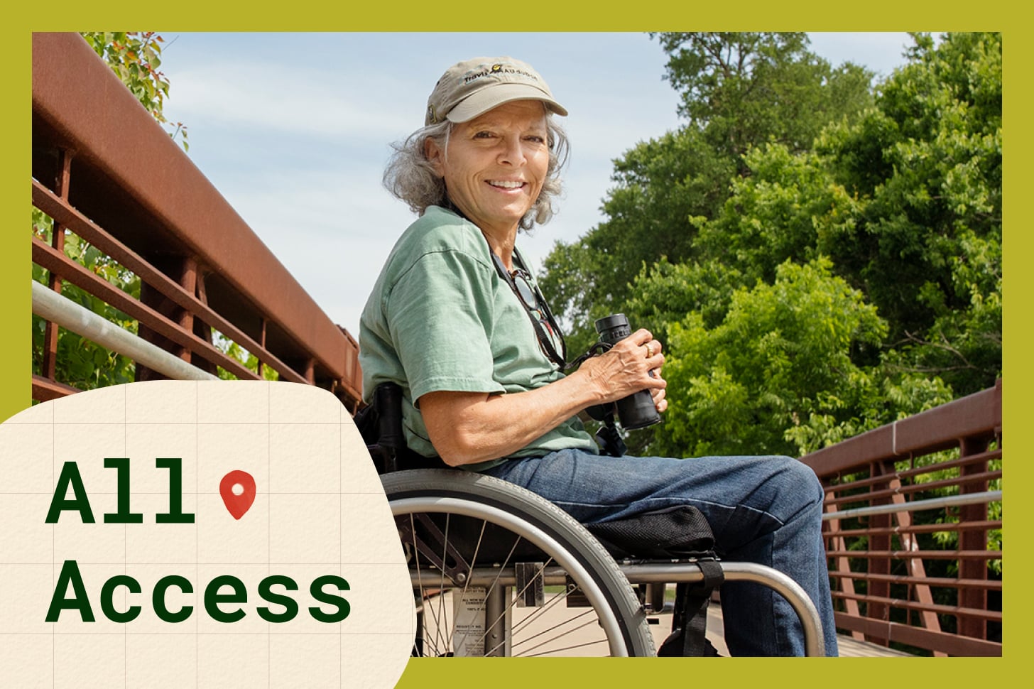Virginia Rose, a fair-skinned woman with grey hair, is a wheelchair-bound birder posing for a photo on a brown bridge while holding black binoculars and wearing a green tshirt, jeans, and a tan hat.