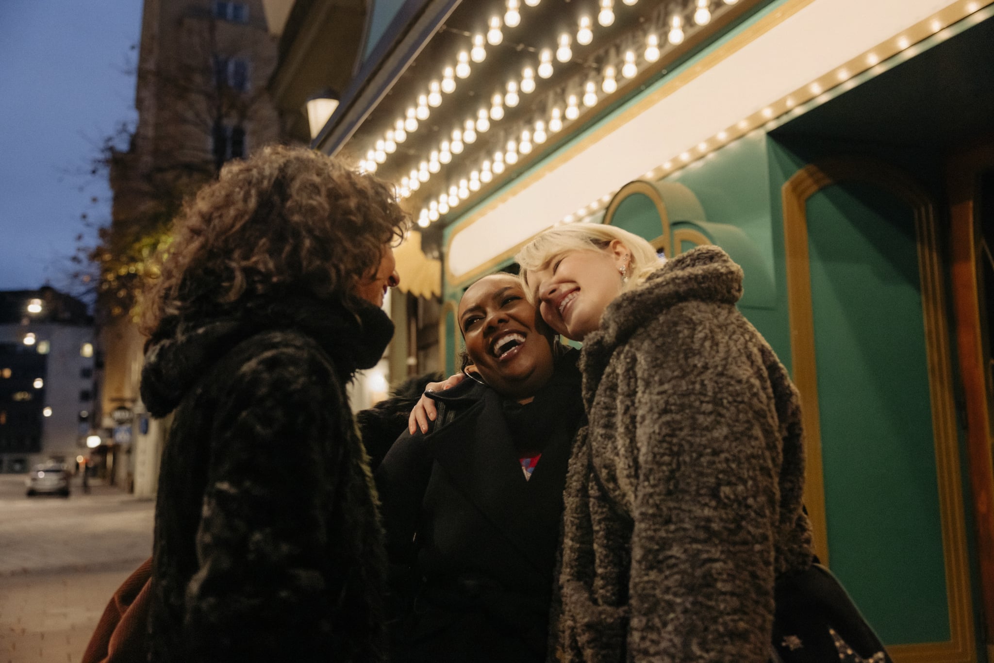 A group of three 20-something women hug and smile outside of a theater