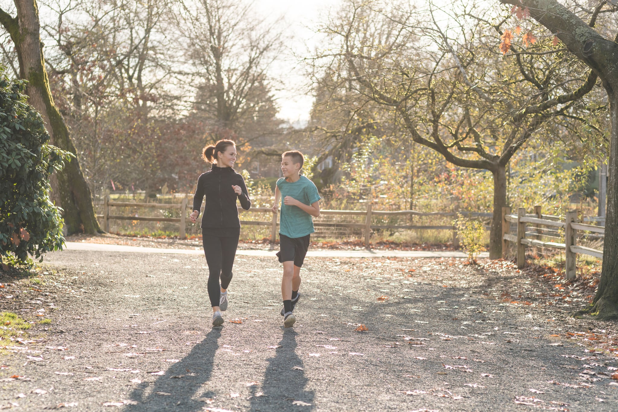 An elementary-age boy goes jogging with his mom in the city park on an autumn morning. They are looking at each other affectionately for a moment as they run. There are leaves scattered on the ground.