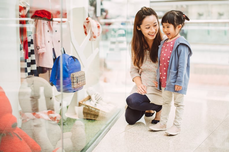 Pretty young mom and lovely little daughter looking at window display for kids wears in a shopping mall joyfully.