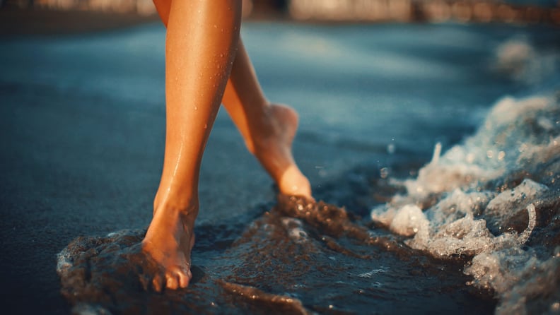 Closeup front view of unrecognizable woman walking on a beach while seawater is splashing on her feet. Shot in a low sunset light.