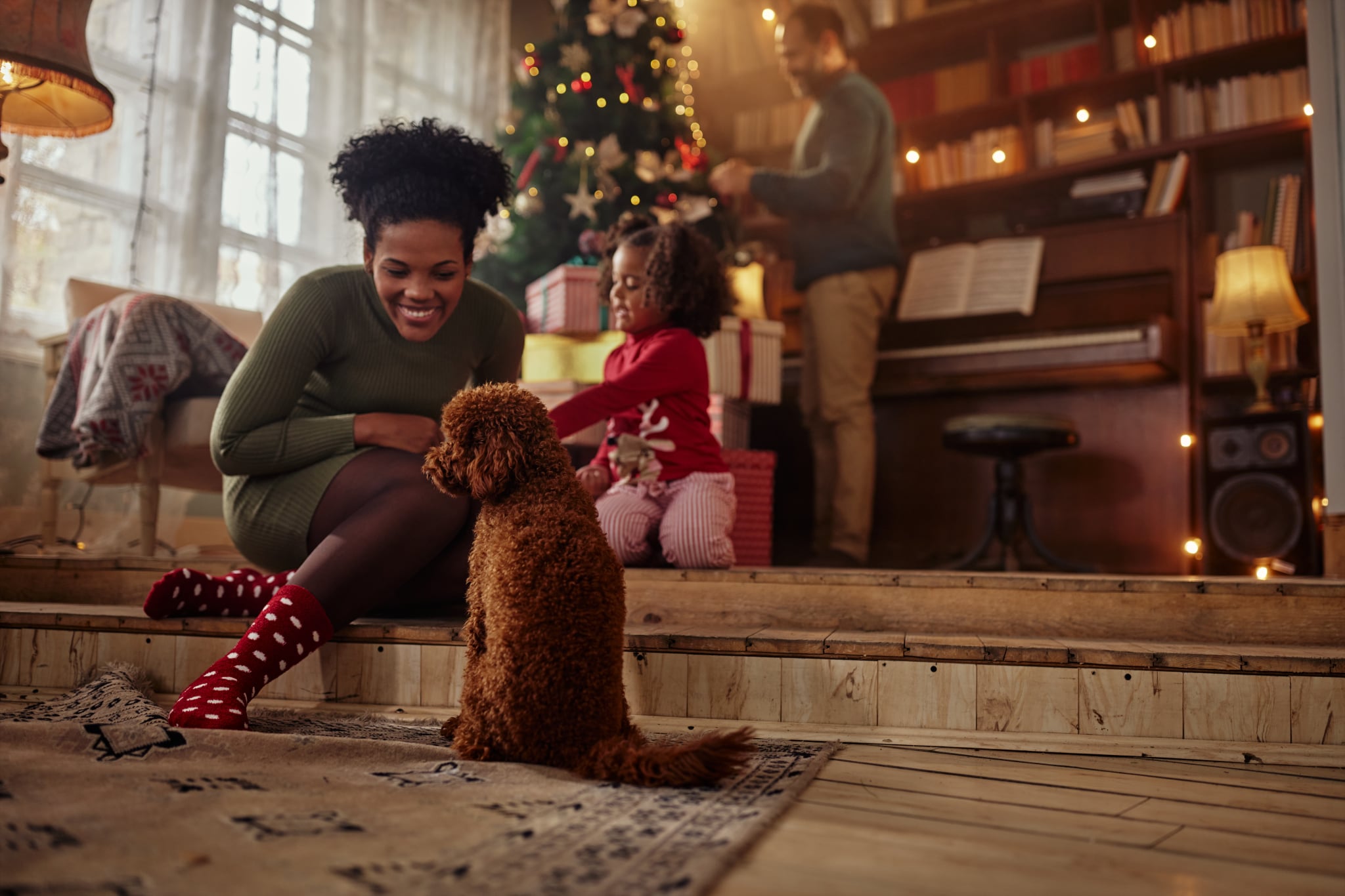 Black woman sitting on the floor and playing with puppy while her daughter unwrapping gifts and husband decorating Christmas tree behind