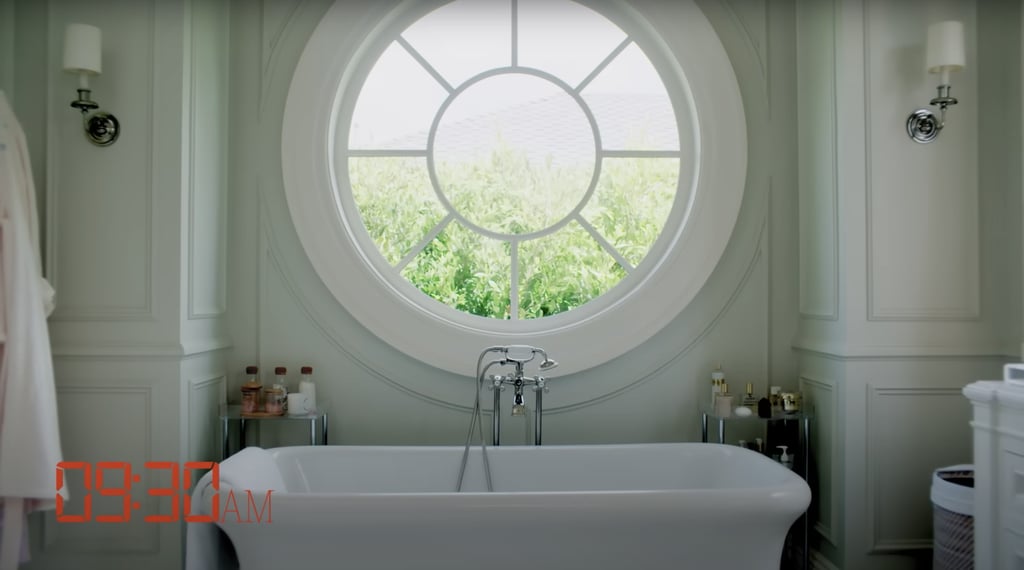 In the bathroom, a large circular window sits above the bathtub, which is flanked by two tables holding candles, perfumes, and other products.