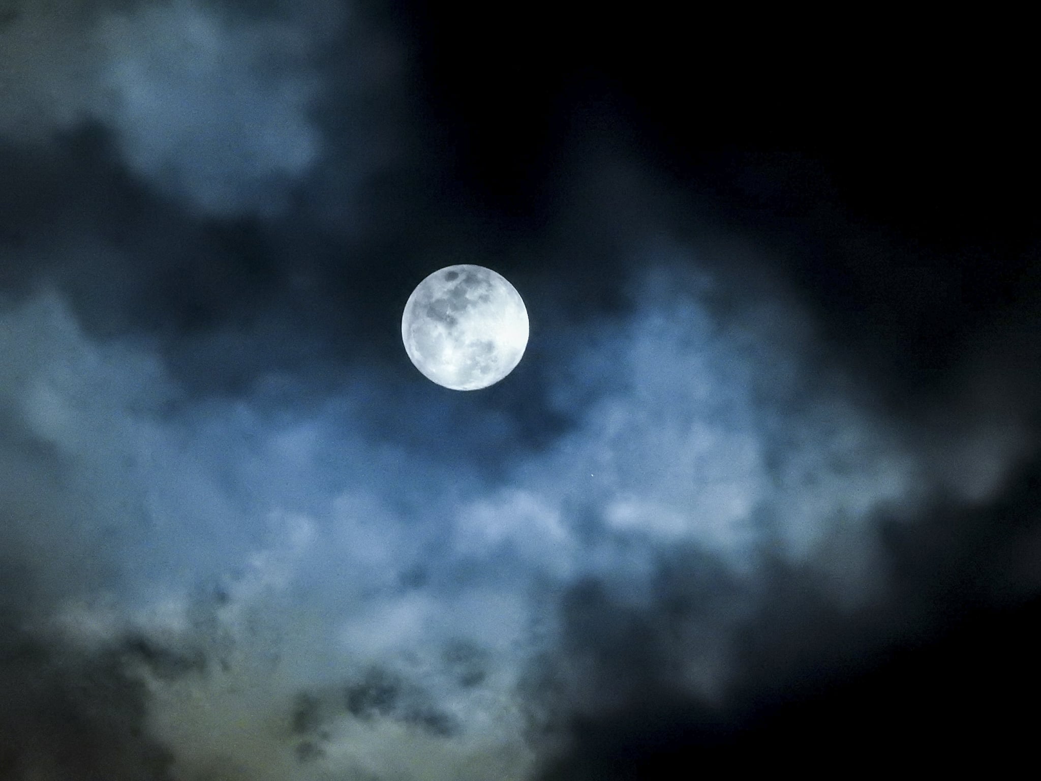 White blue in colour, up close very detailed features of a full moon against a black backdrop of a sky. There are clouds lit up in front of the moon as they pass by.